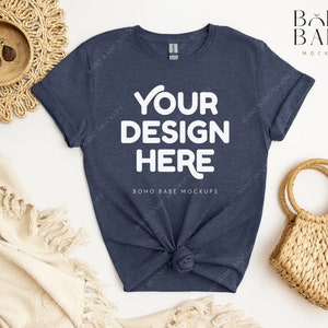 May include: A navy blue t-shirt with the text "YOUR DESIGN HERE" in white. The shirt is knotted at the waist and is surrounded by a woven placemat, earrings, a woven bag, and a cream-colored blanket. The words "BOHO BABE MOCKUPS" are also visible.