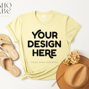May include: A yellow t-shirt with the text "YOUR DESIGN HERE" and "BOHO BABE MOCKUPS" printed on the front. The t-shirt is laid flat on a white surface with a straw hat and sandals.