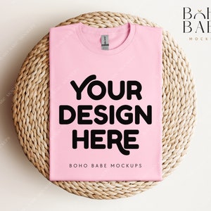 Op de afbeelding: Een roze t-shirt mockup met de tekst "YOUR DESIGN HERE" op een geweven stro mat. De tekst "BOHO BABE MOCKUPS" staat onderaan het shirt.