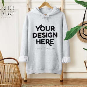 May include: A light gray hoodie with the text "YOUR DESIGN HERE" in black, displayed on a wooden ladder. The sleeves are cuffed to reveal white fabric. The image includes a woven basket, a round woven bag, and greenery.