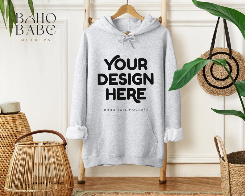 May include: A light gray hoodie with the text "YOUR DESIGN HERE" in black, displayed on a wooden ladder. The sleeves are cuffed to reveal white fabric. The image includes a woven basket, a round woven bag, and greenery.