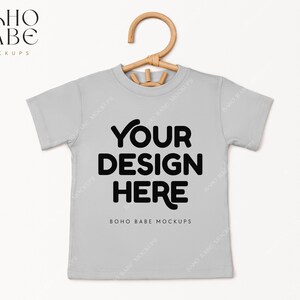 May include: A light gray t-shirt with short sleeves and a hanger. The shirt has the text "YOUR DESIGN HERE" in black and the text "BOHO BABE MOCKUPS" in smaller black text.