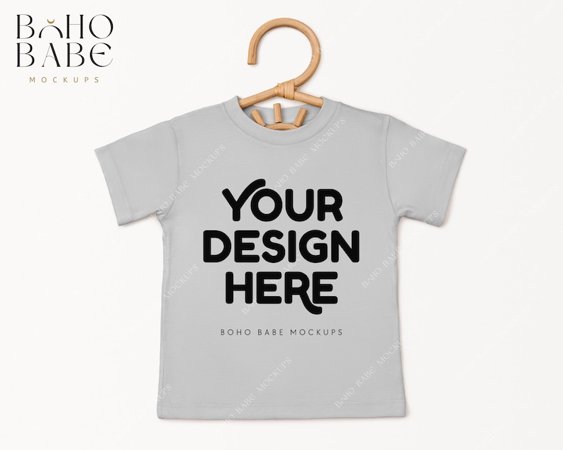 May include: A light gray t-shirt with short sleeves and a hanger. The shirt has the text "YOUR DESIGN HERE" in black and the text "BOHO BABE MOCKUPS" in smaller black text.