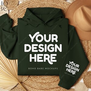 May include: A dark green hooded sweatshirt with the text "YOUR DESIGN HERE" in white. The sweatshirt is laid flat on a woven background with a brown hat and a fan.