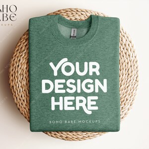 Op de afbeelding: Een gevouwen groen melange t-shirt met de tekst "YOUR DESIGN HERE" in wit op een geweven rieten onderzetter.  BOHO BABE MOCKUPS is gedrukt op de onderkant van het shirt.