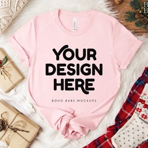 May include: A light pink t-shirt with the text "YOUR DESIGN HERE" in bold black letters. The shirt is displayed on a white fluffy surface, with wrapped gifts and holiday-themed accessories. The shirt is a mock-up for design placement.