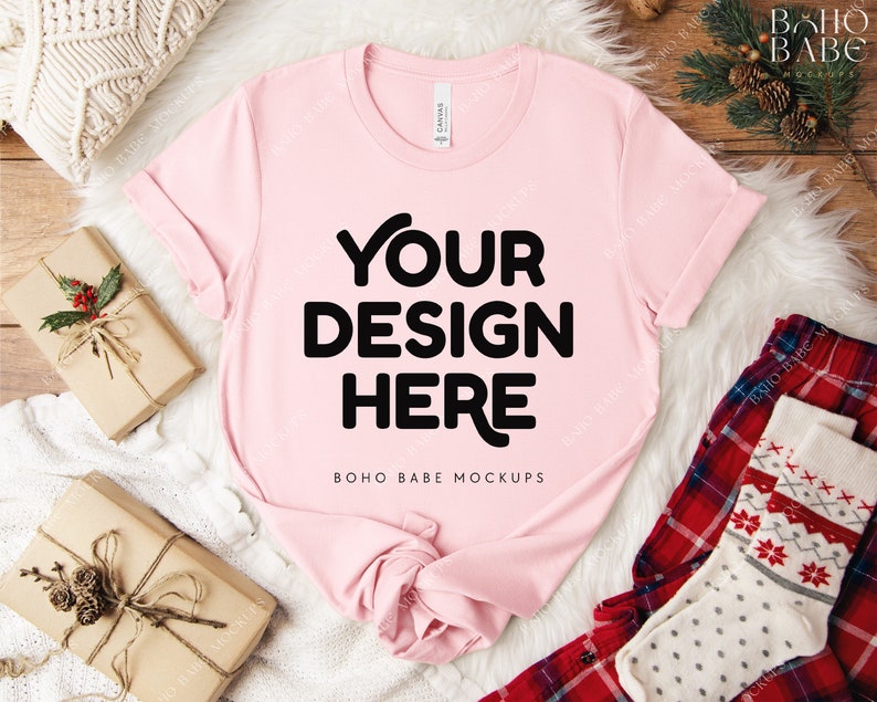 May include: A light pink t-shirt with the text "YOUR DESIGN HERE" in bold black letters. The shirt is displayed on a white fluffy surface, with wrapped gifts and holiday-themed accessories. The shirt is a mock-up for design placement.