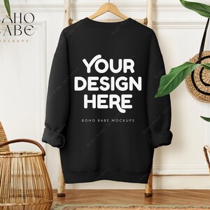 Pode incluir: Mockup de moletom preto com o texto "YOUR DESIGN HERE" na frente. O moletom está pendurado em um cabide de madeira contra uma parede branca.