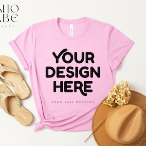 Könnte beinhalten: Ein pinkfarbenes T-Shirt mit dem Text "YOUR DESIGN HERE" und "BOHO BABE MOCKUPS" auf weißem Hintergrund. Das T-Shirt liegt flach mit einem Paar Sandalen und einem braunen Hut daneben.