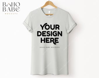 May include: A light gray t-shirt with rolled up sleeves, hanging on a wooden hanger. The shirt has the text "YOUR DESIGN HERE" printed in black on the front. The text "BOHO BABE MOCKUPS" is printed in black below the design.