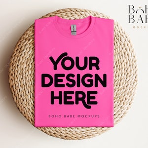 以下が含まれることがあります： ピンクのTシャツのモックアップで、黒い文字で「YOUR DESIGN HERE」と書いてあります。Tシャツは、籐で編まれたマットの上に置かれています。