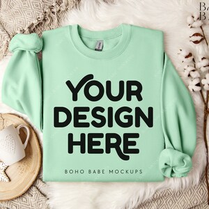 Könnte beinhalten: Ein hellgrüner Pullover mit dem Text "YOUR DESIGN HERE" und "BOHO BABE MOCKUPS" auf der Vorderseite. Der Pullover liegt flach auf einem weißen Kunstfell-Hintergrund.