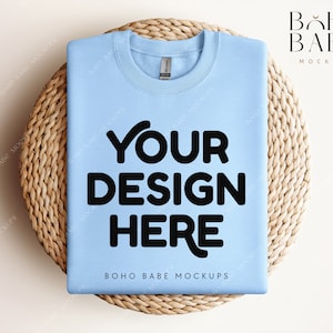 Puede incluir: Una maqueta de sudadera azul claro con el texto "YOUR DESIGN HERE" sobre un posavasos de mimbre tejido. El texto "BOHO BABE MOCKUPS" también es visible en la sudadera.