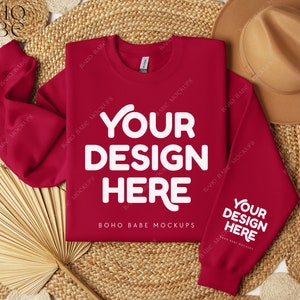May include: Red crewneck sweatshirt with long sleeves, laying flat on a woven surface with a straw hat and a fan. The sweatshirt has the text "YOUR DESIGN HERE" printed on the front and back.