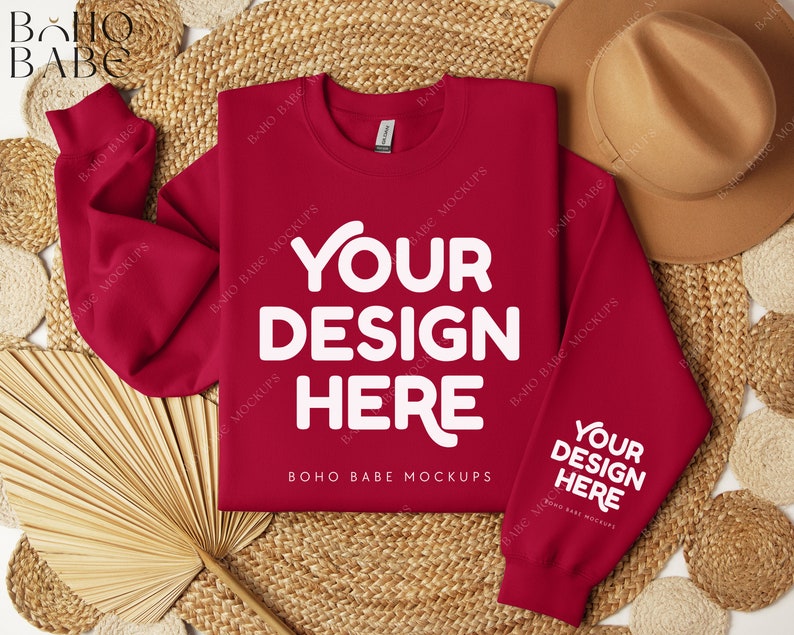 May include: Red crewneck sweatshirt with long sleeves, laying flat on a woven surface with a straw hat and a fan. The sweatshirt has the text "YOUR DESIGN HERE" printed on the front and back.