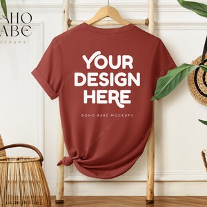 May include: A maroon t-shirt with rolled-up sleeves, hanging on a wooden ladder. The shirt features the text "YOUR DESIGN HERE" in white, with "BOHO BABE MOCKUPS" below. A woven basket and a straw bag are also visible.
