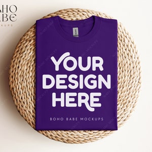 May include: Purple t-shirt mockup with the text "YOUR DESIGN HERE" on a woven straw mat.  BOHO BABE MOCKUPS is printed on the bottom of the shirt.
