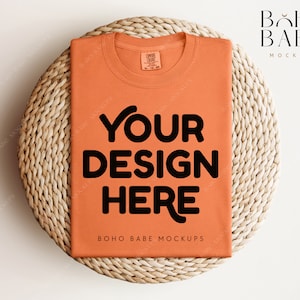 May include: A flat lay mockup of a burnt orange t-shirt with the text "YOUR DESIGN HERE" on a woven wicker surface. The text "BOHO BABE MOCKUPS" is printed on the bottom of the t-shirt.