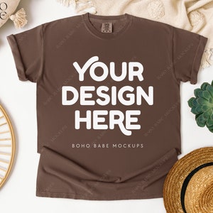 May include: A brown t-shirt with the text "YOUR DESIGN HERE" in white. The shirt is displayed on a white surface with a straw hat, a succulent plant, and a woven circular decoration. The text "BOHO BABE MOCKUPS" is also visible.
