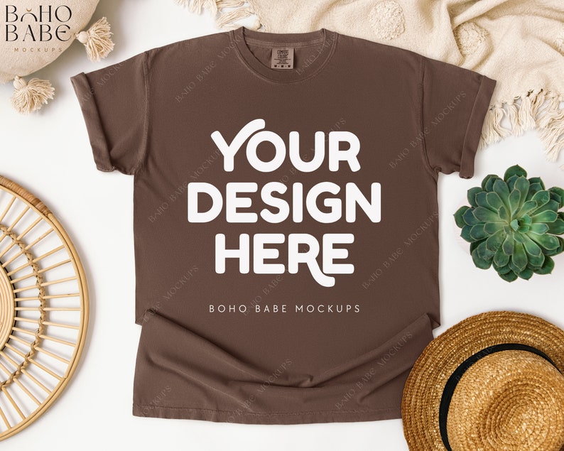 May include: A brown t-shirt with the text "YOUR DESIGN HERE" in white. The shirt is displayed on a white surface with a straw hat, a succulent plant, and a woven circular decoration. The text "BOHO BABE MOCKUPS" is also visible.