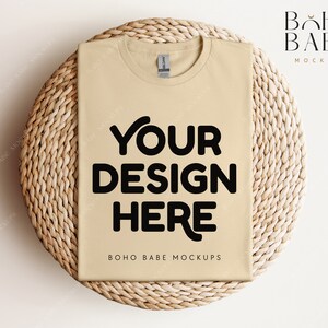 Op de afbeelding: Een beige t-shirt mockup op een geweven rieten oppervlak. De tekst "YOUR DESIGN HERE" is in zwart op de voorkant van het shirt gedrukt. De tekst "BOHO BABE MOCKUPS" is onder het ontwerp gedruckt.