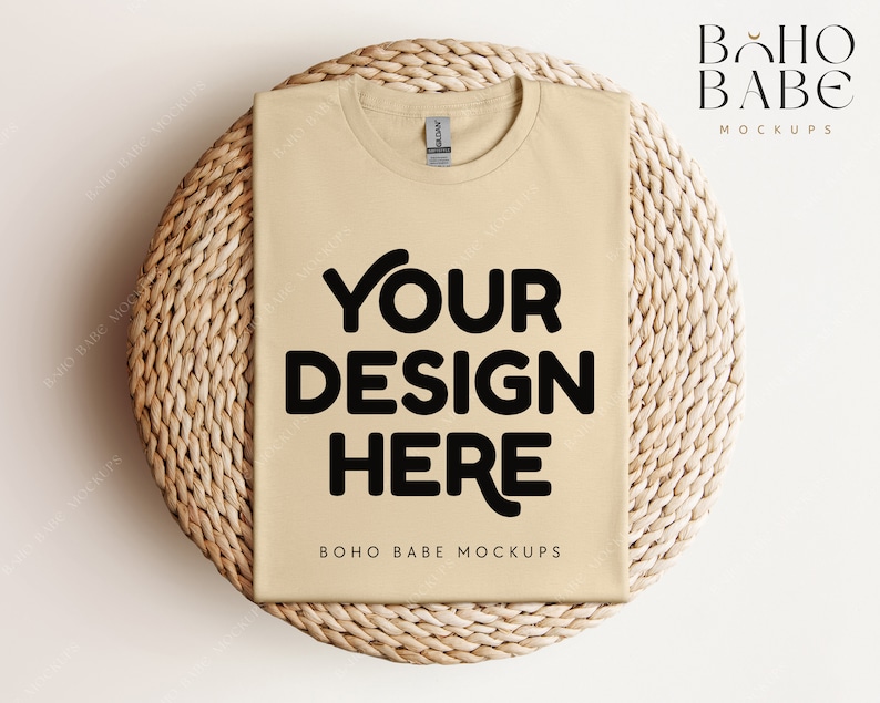 Op de afbeelding: Een beige t-shirt mockup op een geweven rieten oppervlak. De tekst "YOUR DESIGN HERE" is in zwart op de voorkant van het shirt gedrukt. De tekst "BOHO BABE MOCKUPS" is onder het ontwerp gedruckt.