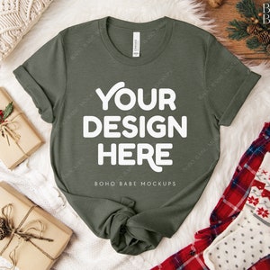May include: An olive green t-shirt with the text "YOUR DESIGN HERE" in white. The shirt is displayed on a white faux fur surface, with wrapped gifts and holiday-themed socks and pajamas. The shirt is a mock-up.