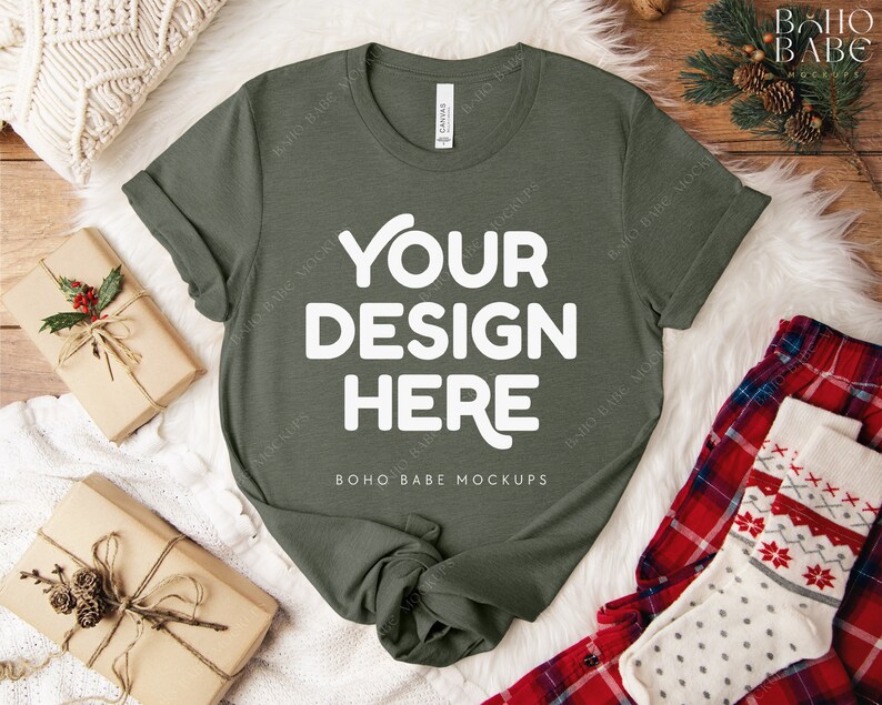 May include: An olive green t-shirt with the text "YOUR DESIGN HERE" in white. The shirt is displayed on a white faux fur surface, with wrapped gifts and holiday-themed socks and pajamas. The shirt is a mock-up.