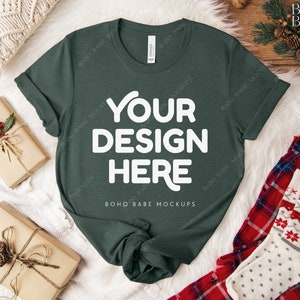May include: A dark green t-shirt with the text "YOUR DESIGN HERE" in white. The t-shirt is laying on a white fuzzy surface with a brown wooden surface in the background. There are two wrapped gifts and a pair of red and white plaid pajamas with white socks on the surface.