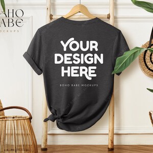 Op de afbeelding: Een donkergrijs T-shirt met de tekst "YOUR DESIGN HERE" en "BOHO BABE MOCKUPS" gedrukt op de achterkant. Het T-shirt hangt aan een houten ladder tegen een witte muur.