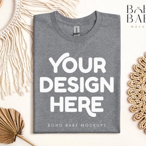 May include: A flat lay mockup of a heather gray t-shirt with the text "YOUR DESIGN HERE" printed in white. The t-shirt is surrounded by natural elements, including a woven macrame wall hanging, a palm leaf, and a woven straw placemat.