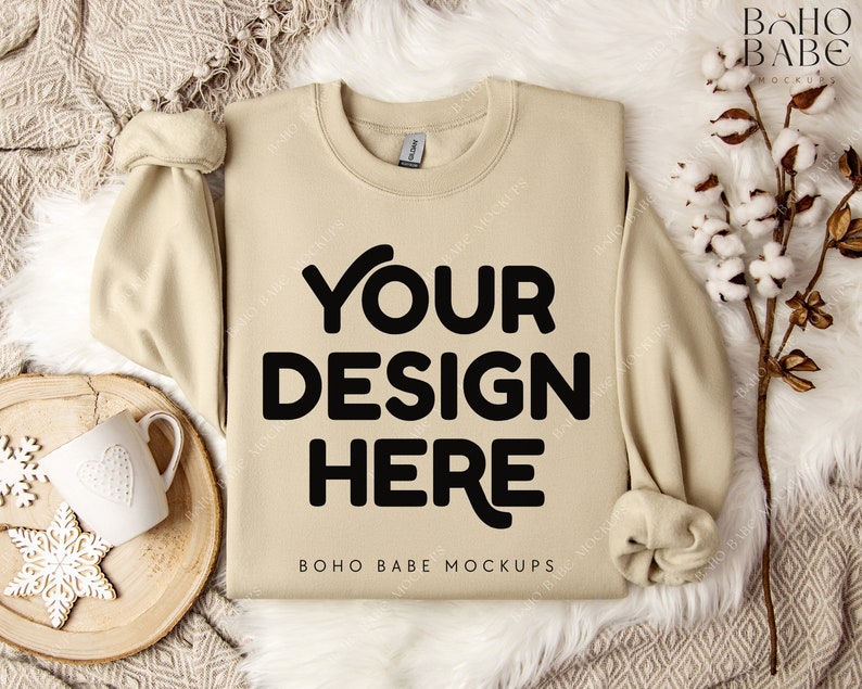 May include: A beige sweatshirt with the text "YOUR DESIGN HERE" in black. The sweatshirt is displayed on a white fur-like surface, with a wooden tray, a white mug, and decorative snowflakes. The image is a mockup.