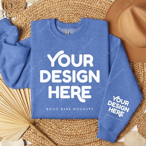 May include: A light blue sweatshirt with the text "YOUR DESIGN HERE" in white on the front and the text "BOHO BABE MOCKUPS" in white below. The sweatshirt is on a woven straw background with a brown hat and a fan.
