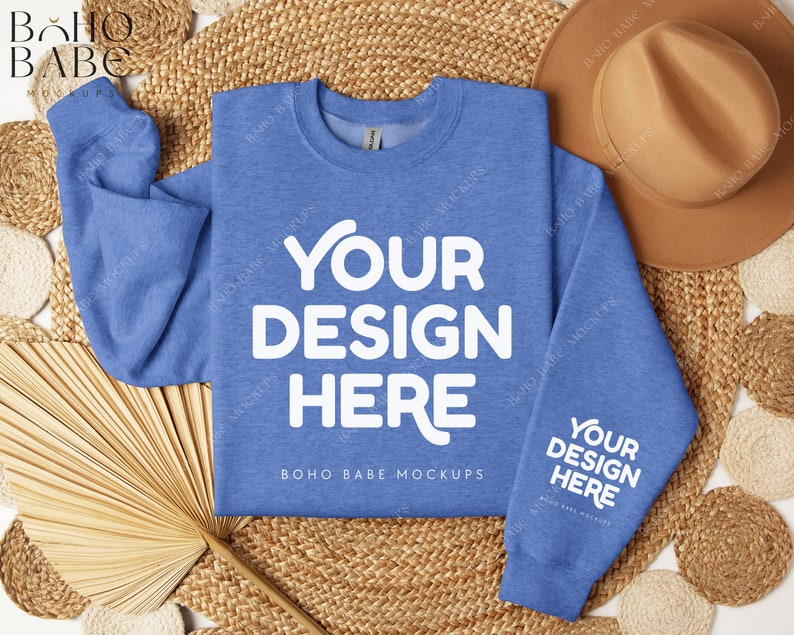 May include: A light blue sweatshirt with the text "YOUR DESIGN HERE" in white on the front and the text "BOHO BABE MOCKUPS" in white below. The sweatshirt is on a woven straw background with a brown hat and a fan.