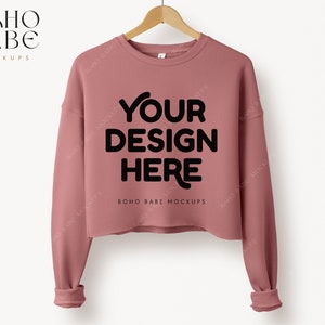 以下が含まれることがあります： 長袖のピンク色のクロップドスウェットシャツ。スウェットシャツには、「YOUR DESIGN HERE」と「BOHO BABE MOCKUPS」の文字が前面に印刷されています。