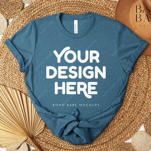 Peut inclure: Un t-shirt bleu sarcelle avec le texte "YOUR DESIGN HERE" en blanc, pr&eacute;sent&eacute; sur un tapis tiss&eacute; avec des &eacute;l&eacute;ments d&eacute;coratifs. Le t-shirt est une maquette, parfaite pour pr&eacute;senter des cr&eacute;ations.