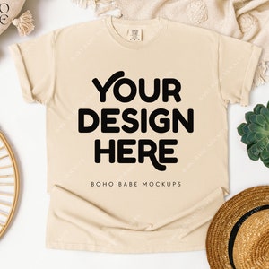 May include: Cream-colored t-shirt with rolled-up sleeves, featuring the text "YOUR DESIGN HERE" in bold black letters. The mock-up includes a straw hat, a succulent plant, and a decorative woven plate.