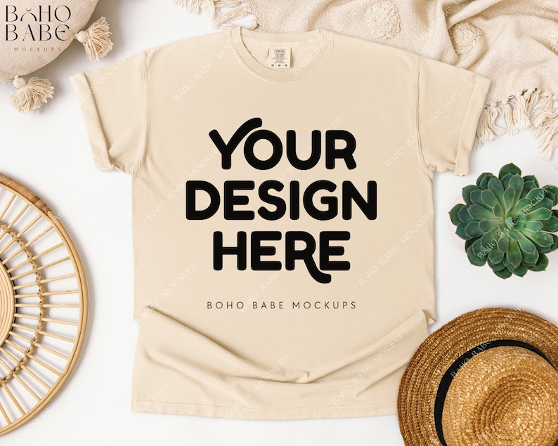 May include: Cream-colored t-shirt with rolled-up sleeves, featuring the text "YOUR DESIGN HERE" in bold black letters. The mock-up includes a straw hat, a succulent plant, and a decorative woven plate.