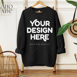 May include: Black sweatshirt mockup with the text "YOUR DESIGN HERE" on the front. The sweatshirt is hanging on a wooden hanger against a white wall.