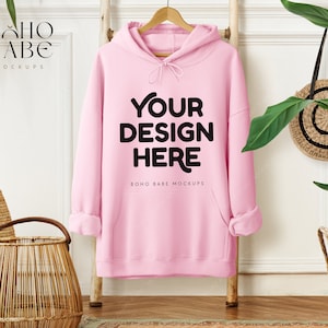May include: A light pink hoodie with the text "YOUR DESIGN HERE" in black. The hoodie is hanging on a wooden hanger against a white wall. The words "BOHO BABE MOCKUPS" are printed on the top left and bottom center of the image.