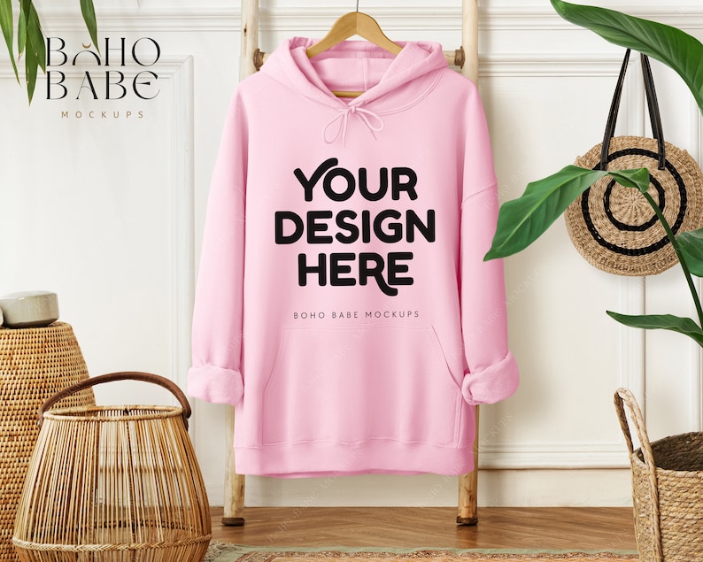 May include: A light pink hoodie with the text "YOUR DESIGN HERE" in black. The hoodie is hanging on a wooden hanger against a white wall. The words "BOHO BABE MOCKUPS" are printed on the top left and bottom center of the image.