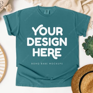 Puede incluir: Una maqueta de camiseta azul turquesa con el texto "YOUR DESIGN HERE" en blanco. La camiseta está colocada sobre una superficie blanca con un sombrero de paja, una planta suculenta y una cesta de mimbre.