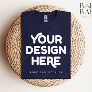 Op de afbeelding: Een mockup van een marineblauw t-shirt met de tekst "YOUR DESIGN HERE" in wit op een geweven strooien mat. De tekst "BOHO BABE MOCKUPS" is ook zichtbaar op het t-shirt.