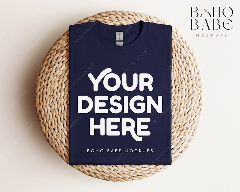 Op de afbeelding: Een mockup van een marineblauw t-shirt met de tekst "YOUR DESIGN HERE" in wit op een geweven strooien mat. De tekst "BOHO BABE MOCKUPS" is ook zichtbaar op het t-shirt.