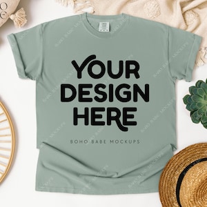 May include: A sage green t-shirt with the text "YOUR DESIGN HERE" in bold black letters. The shirt is laid flat on a white surface, with a straw hat, succulent, and decorative elements around it. The text "BOHO BABE MOCKUPS" is also visible.