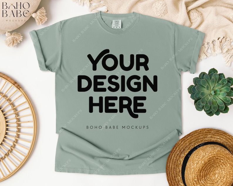 May include: A sage green t-shirt with the text "YOUR DESIGN HERE" in bold black letters. The shirt is laid flat on a white surface, with a straw hat, succulent, and decorative elements around it. The text "BOHO BABE MOCKUPS" is also visible.