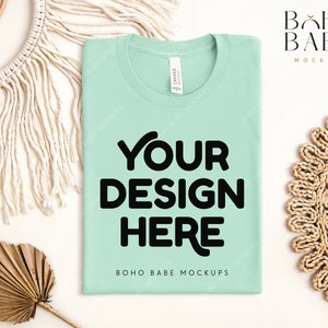 May include: A light green t-shirt mockup with the text "YOUR DESIGN HERE" in black. The t-shirt is lying flat on a white surface with a woven straw mat and a palm leaf to the right and a macrame wall hanging to the left. The text "BOHO BABE MOCKUPS" is printed below the design.
