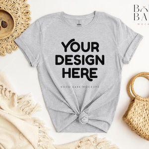 May include: A gray t-shirt with the text "YOUR DESIGN HERE" and "BOHO BABE MOCKUPS" printed on it. The t-shirt is laid flat on a white surface with a woven basket and a blanket.