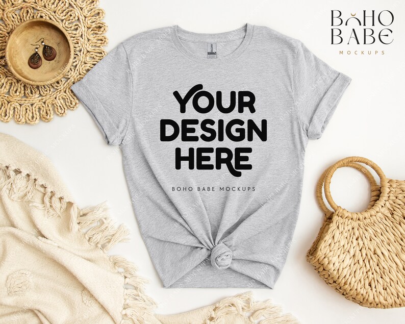 May include: A gray t-shirt with the text "YOUR DESIGN HERE" and "BOHO BABE MOCKUPS" printed on it. The t-shirt is laid flat on a white surface with a woven basket and a blanket.