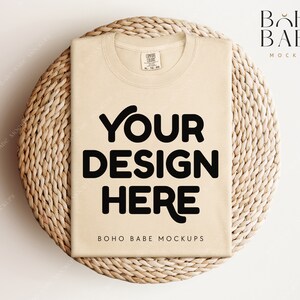 May include: A beige t-shirt mockup on a woven straw mat. The t-shirt has the text "YOUR DESIGN HERE" printed on the front. The text "BOHO BABE MOCKUPS" is printed below the design.